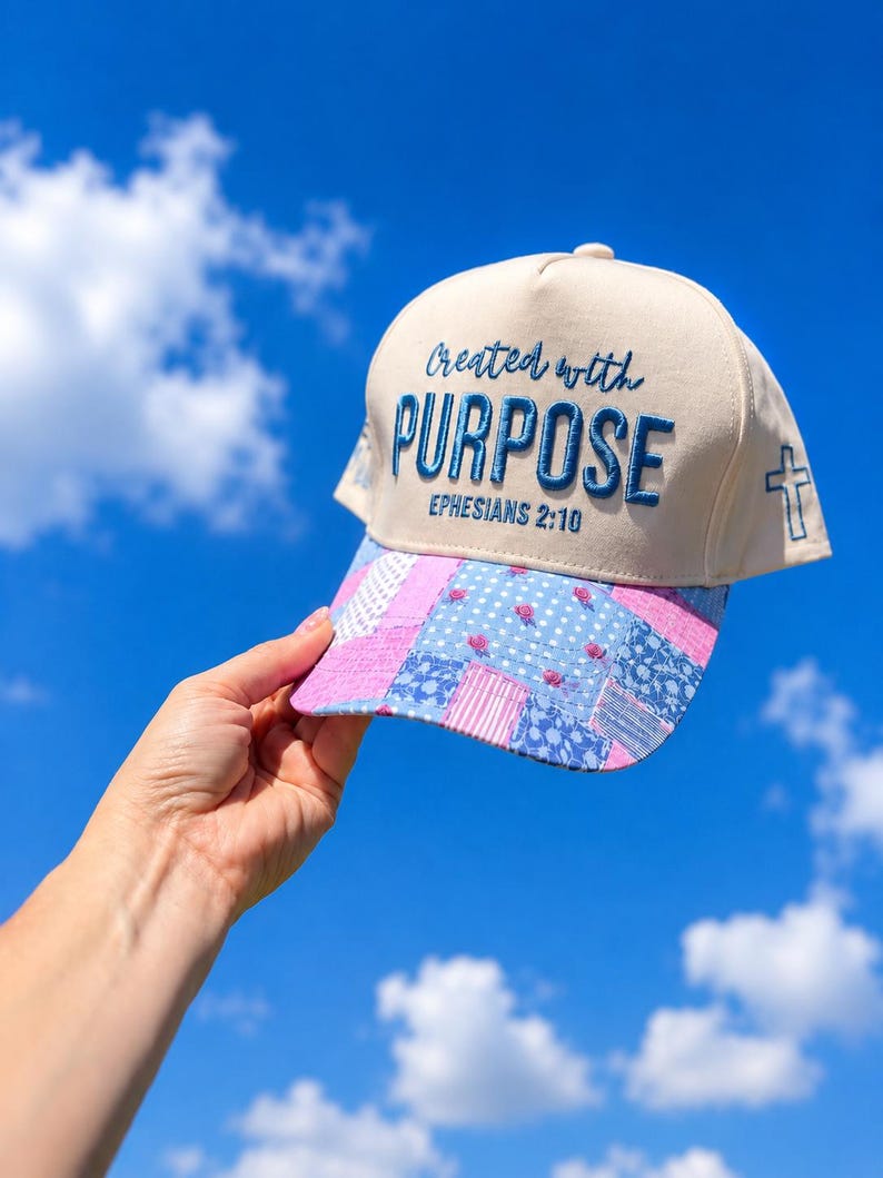 K&ouml;nnte beinhalten: Eine beige Baseballkappe mit blauer Stickerei, die den Text Created with PURPOSE EPHESIANS 2:10 zeigt. Der Schirm ist im Patchwork-Stil mit blauen und rosafarbenen Blumen- und Streifenmustern gestaltet und an der Seite befindet sich ein Kreuz.
