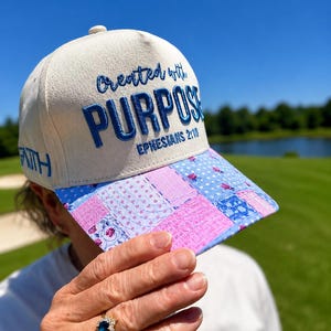 K&ouml;nnte beinhalten: Eine cremefarbene Baseballkappe mit blauem Sticktext, der Created with PURPOSE EPHESIANS 2:10 lautet. An der Seite der Kappe ist das Wort FAITH in blauer Stickerei zu sehen. Der Schirm zeigt ein blau-rosa Blumenmuster im Patchwork-Stil.