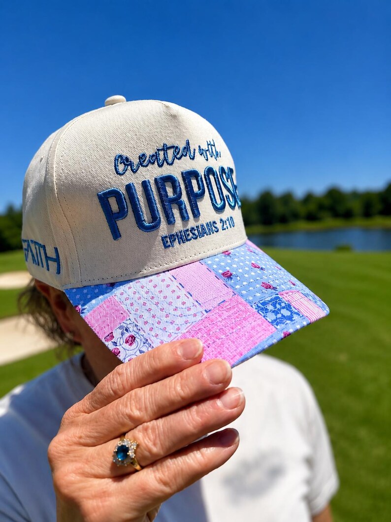 K&ouml;nnte beinhalten: Eine cremefarbene Baseballkappe mit blauem Sticktext, der Created with PURPOSE EPHESIANS 2:10 lautet. An der Seite der Kappe ist das Wort FAITH in blauer Stickerei zu sehen. Der Schirm zeigt ein blau-rosa Blumenmuster im Patchwork-Stil.