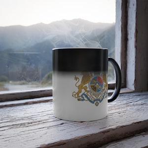 May include: A colour-changing mug, black at the top and white at the bottom, displaying a detailed coat of arms with lions and a shield. The mug is set against a window with a mountain view.