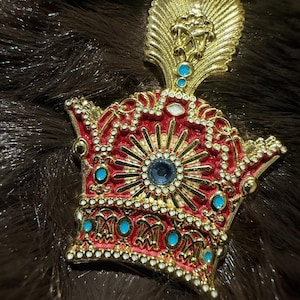 May include: A gold-tone metal brooch shaped like a crown, featuring a red enamel base, clear rhinestones along the edges, and small turquoise-coloured circular accents. The centre displays a sunburst design with a single blue stone at its core.