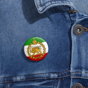 Puede incluir: Un botón redondo con la bandera de Irán. El botón tiene un esquema de color verde, blanco y rojo con un emblema de león y corona dorados en el centro. El botón está sujeto a una chaqueta vaquera azul.