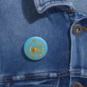 May include: A round, light blue button pin featuring a gold lion and sun emblem within a gold wreath. The pin is set against a blue denim jacket.