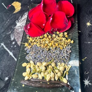 May include: A rectangular black tray decorated with white celestial patterns, holding layers of dried red rose petals, yellow chamomile flowers, purple lavender buds, white jasmine buds, and two dark brown vanilla beans arranged in horizontal rows.