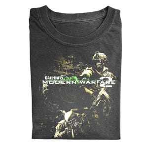 May include: Dark gray t-shirt featuring a graphic from the video game "Call of Duty: Modern Warfare 2." The design includes soldiers, military vehicles, and the game's logo in white and green. The shirt is folded.