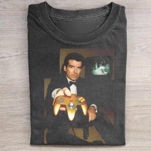 May include: Dark gray t-shirt featuring a graphic of a man in a tuxedo holding a gold-colored video game controller. The image includes a television screen showing a video game scene. The shirt is folded.