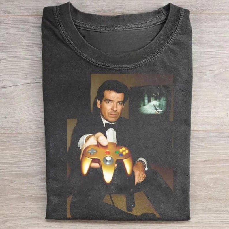 May include: Dark gray t-shirt featuring a graphic of a man in a tuxedo holding a gold-colored video game controller. The image includes a television screen showing a video game scene. The shirt is folded.