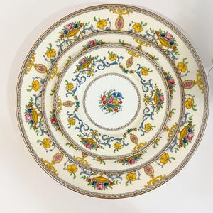 May include: A set of three cream-colored porcelain plates with a floral design. The plates feature a gold rim and are decorated with yellow, blue, pink, and purple flowers and baskets. The plates are arranged in a nested configuration.