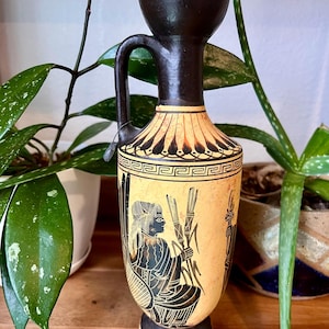 May include: A ceramic lekythos vase featuring a black-figure design on a tan background. The vessel has a tall neck, a single handle, and a flared rim. The body displays a seated figure holding stalks, with decorative geometric patterns above.