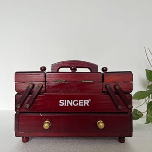 Puede incluir: Una caja de costura Singer de madera roja oscura con asa, cajones y compartimentos. La caja tiene una tapa con bisagras y está adornada con pomos de latón. El logotipo de Singer se muestra de forma destacada en la parte delantera.