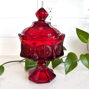 Może przedstawiać: Rubinowoczerwony, zakryty pojemnik na cukierki z podstawą. Szklane naczynie posiada wytłoczone okrągłe wzory i fasetowany finial na pokrywie. Naczynie jest prezentowane na białej powierzchni z zielonymi liśćmi w tle.