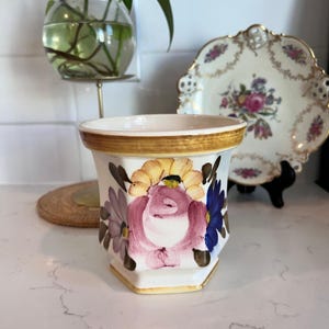 Op de afbeelding: Een witte keramische bloempot met een gouden rand en een handgeschilderd bloemenontwerp met roze rozen, gele en blauwe bloemen. De zeshoekige bloempot staat op een wit oppervlak.