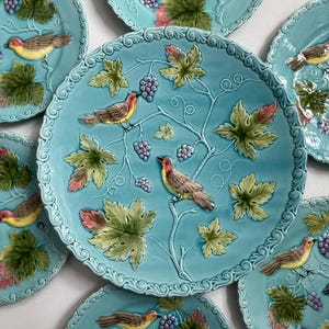 May include: A collection of turquoise ceramic plates with a raised relief design. Each plate features a bird perched on a vine with grapes and leaves. The plates have a scalloped edge and are arranged on a white surface.