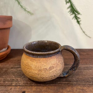 May include: A handmade ceramic mug with a rounded body and a sturdy handle. The mug features a speckled, earthy brown and tan color palette, with a darker brown rim and handle. The mug is designed for hot beverages.