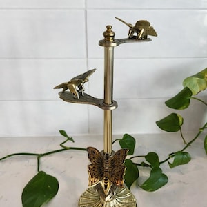 May include: A brass-toned, tiered candle holder with butterfly and bird accents. The base features a butterfly design, while the upper tiers hold small bird figures. The candle holder is a decorative item, likely for home decor.