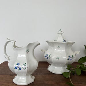 May include: White ceramic creamer and covered sugar bowl set. Both pieces feature a floral pattern with blue flowers and green leaves. The creamer has a handle and spout, while the sugar bowl has handles and a lid.