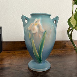 May include: A light blue ceramic vase with a floral design. The vase features a raised iris flower in shades of cream, yellow, and green. It has two handles and a pedestal base. The vase is approximately 6 inches tall.