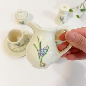 Puede incluir: Un juego de té en miniatura de cerámica color crema con un diseño floral. El juego incluye una jarra de crema, azucarero, taza, platillo y un pequeño agitador. Cada pieza está decorada con un motivo floral morado y verde.