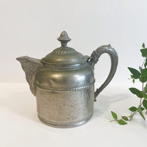 Könnte beinhalten: Antike silberfarbene Teekanne mit dekorativem Griff und Ausguss. Die Teekanne hat einen gewölbten Deckel mit einem kleinen Knauf. Der Griff weist ein verziertes Design auf. Die Teekanne ist aus Metall und hat ein Vintage-Aussehen.