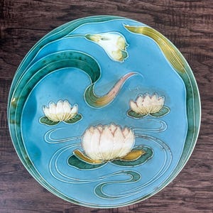 May include: A round, ceramic plate with a light blue background and a water lily design. The plate features white water lilies, green lily pads, and curved lines suggesting water. The rim is a darker teal green.