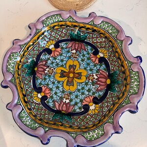 May include: A hand-painted ceramic decorative plate with a scalloped edge. The plate features a vibrant floral design in shades of blue, yellow, pink, and green, with a dark blue border and a lavender rim. The plate's diameter is approximately 12 inches.