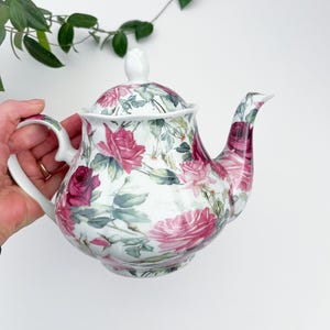 May include: White ceramic teapot with a lid, handle, and spout. The teapot is decorated with a floral pattern of pink roses and green leaves. The teapot is a classic design, perfect for serving tea.