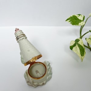 Può includere: Un portagioie a forma di faro in ceramica bianca con dettagli color oro. La parte superiore del faro è rosa e bianca e la scatola è aperta, rivelando un piccolo disegno di faro all'interno. La scatola è su una superficie bianca con una pianta verde e bianca sullo sfondo.