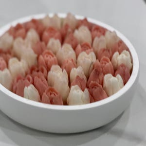 Puede incluir: Un cuenco de cerámica blanco lleno de numerosos dulces con forma de tulipán. Los dulces son una mezcla de rosa claro y blanco, dispuestos para parecerse a un campo de tulipanes. El cuenco está sobre una superficie blanca.