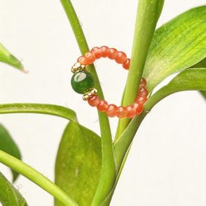 Puede incluir: Un anillo de cuentas con una piedra verde central y cuentas naranjas. El anillo se encuentra sobre un fondo de tallos y hojas verdes, con un fondo blanco. El diseño del anillo incorpora detalles dorados.