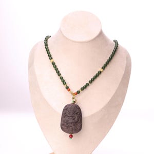 May include: A dark brown pendant necklace featuring a carved dragon design. The necklace has green beads and gold accents. The pendant is suspended from the beaded chain. Displayed on a mannequin.