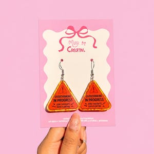 Op de afbeelding: Een paar oranje en gele oorbellen met een waarschuwingsbordontwerp. De oorbellen hebben de tekst "OVERTHINKING IN PROGRESS" en "ZERO THOUGHTS, 100% FLOATING". De oorbellen zitten op een roze kaart met "Mar of Creativ."