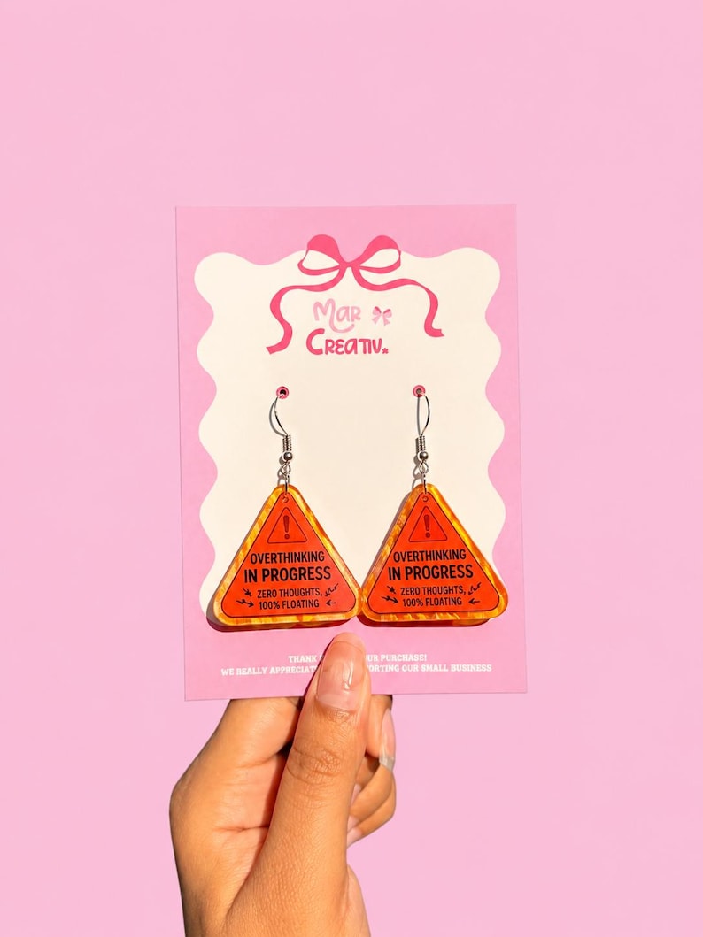 Op de afbeelding: Een paar oranje en gele oorbellen met een waarschuwingsbordontwerp. De oorbellen hebben de tekst "OVERTHINKING IN PROGRESS" en "ZERO THOUGHTS, 100% FLOATING". De oorbellen zitten op een roze kaart met "Mar of Creativ."