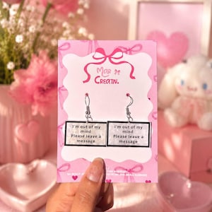 May include: A pair of silver-tone earrings with rectangular pendants. Each pendant has the text "I'm out of my mind Please leave a message" in a black frame. The earrings are displayed on a pink card with the text "Mar de Creativ."