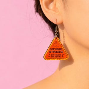 Op de afbeelding: Een enkele oranje en zwarte oorbel in de vorm van een waarschuwingstriangel. De oorbel heeft de tekst "OVERTHINKING IN PROGRESS" en "ZERO THOUGHTS, 100% FLOATING". De oorbel hangt aan een zilverkleurige haak.