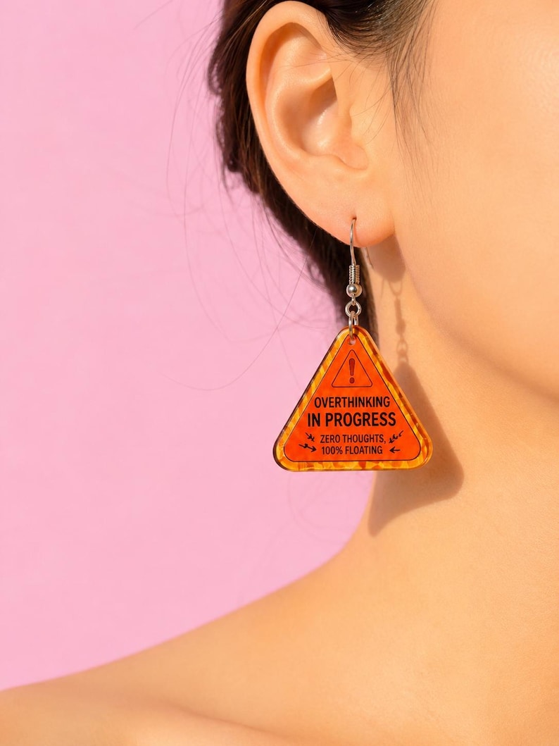 Op de afbeelding: Een enkele oranje en zwarte oorbel in de vorm van een waarschuwingstriangel. De oorbel heeft de tekst "OVERTHINKING IN PROGRESS" en "ZERO THOUGHTS, 100% FLOATING". De oorbel hangt aan een zilverkleurige haak.