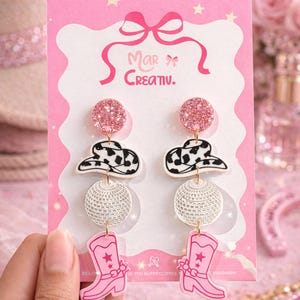 May include: Pink and white novelty earrings with a cowboy theme. The earrings feature a pink glitter ball, a black and white cow print cowboy hat, a disco ball, and a pink cowboy boot. The earrings are displayed on a pink card with the text "Mar Creativ."