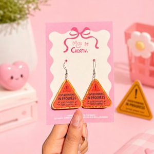 May include: A pair of dangle earrings featuring a warning sign design with the text "OVERTHINKING IN PROGRESS." The earrings are orange and yellow with silver hooks. The earrings are displayed on a pink card.