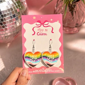 May include: A pair of heart-shaped earrings with a rainbow design and the text "SAFE WITH ME". The earrings are on a pink card with the brand name "Mar Creativ.". The card also includes the text "THANK YOU FOR YOUR PURCHASED I REALLY APPRECIATE YOU SUPPORTING OUR SMALL BUSINESS".