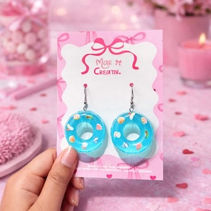 May include: A pair of blue resin donut earrings with colorful sprinkles and seashell accents. The earrings are displayed on a pink card with the text "Mar Creativ." and a bow design. The background is a soft pink.