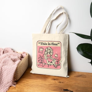 Könnte beinhalten: Beige Tragetasche mit rosa und weißer Grafik. Die Grafik zeigt "This is fine" und zeigt eine Frau in einem rosa Kleid, die in einem Stuhl sitzt. Eine Lampe, eine Pflanze und ein Bilderrahmen sind ebenfalls vorhanden.