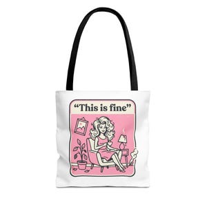 Könnte beinhalten: Weiße Tragetasche mit schwarzen Griffen. Auf der Tasche befindet sich eine rosa Grafik einer Frau, die in einem Stuhl sitzt und eine Tasse hält, mit dem Text "This is fine" darüber. Eine Lampe, eine Pflanze und ein Bilderrahmen sind ebenfalls in der Grafik.