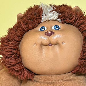 May include: Close-up of a vintage Cabbage Patch Kids doll with a lion-like appearance. The doll has a tan face, blue eyes, and a small pink nose. It features a brown, shaggy mane and is wearing a brown outfit.