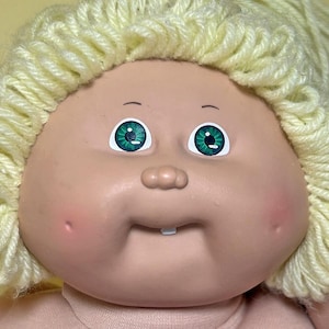 May include: Close-up of a vintage doll's face. The doll has a fair complexion, rosy cheeks, and a small, upturned nose. Its eyes are a vibrant green with black pupils. The doll has a small mouth with a single tooth and light yellow yarn hair.