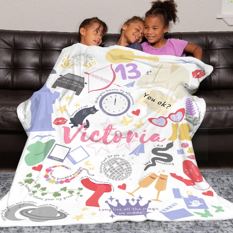 May include: A white blanket with a collage of illustrations and text, including the name "Victoria" in pink. The design features a clock, sunglasses, a cat, a car, and the number "13". The blanket is draped over a dark brown sofa.
