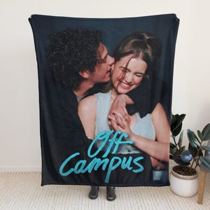 Pu&ograve; includere: Una coperta rettangolare con una stampa fotografica di due persone che si abbracciano. La persona a sinistra indossa una giacca nera, mentre quella a destra indossa un top azzurro. Il testo Off Campus &egrave; stampato in un colore blu brillante.