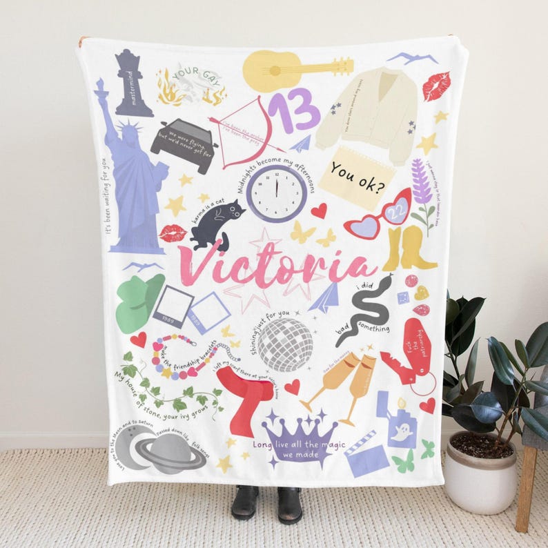 May include: White blanket featuring a collage of illustrations, including a guitar, a car, a cat, a clock, and the name "Victoria" in pink. The blanket has various text elements and colourful graphics.