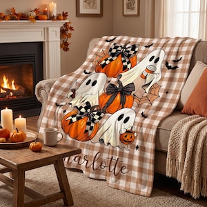 May include: A beige and white buffalo plaid throw blanket featuring a printed design of three white ghosts, orange pumpkins with black and white checkered bows, spiderwebs, and small black bats. The name Charlotte is printed in brown script.