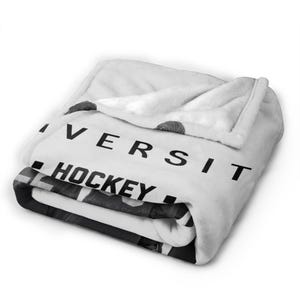 Pu&ograve; includere: Una coperta in pile bianca piegata con una stampa grafica in bianco e nero. Il testo "IVERSIT" &egrave; visibile in grandi lettere maiuscole sopra la parola "HOCKEY" in grassetto nero. Il tessuto ha una consistenza morbida e felpata con pieghe.