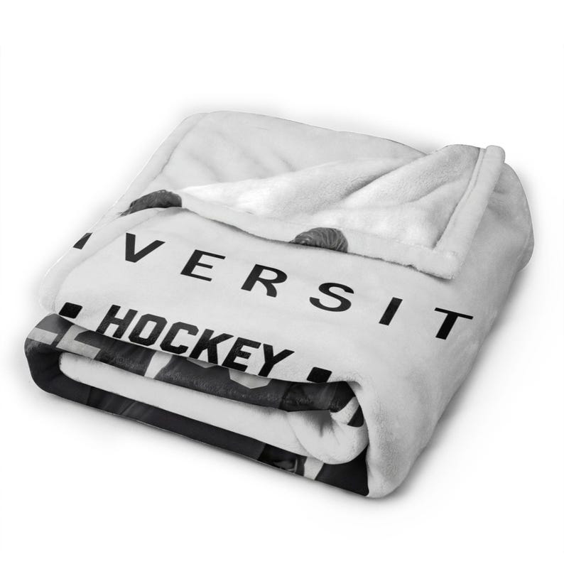 Pu&ograve; includere: Una coperta in pile bianca piegata con una stampa grafica in bianco e nero. Il testo "IVERSIT" &egrave; visibile in grandi lettere maiuscole sopra la parola "HOCKEY" in grassetto nero. Il tessuto ha una consistenza morbida e felpata con pieghe.