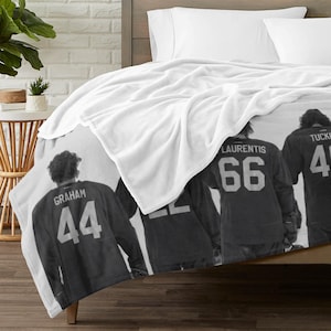 Pu&ograve; includere: Una coperta in pile bianca con una fotografia in bianco e nero di quattro giocatori di hockey visti da dietro. I giocatori indossano maglie con i nomi Graham, Laurentis e Tucker, insieme ai numeri 44, 22, 66 e 41 stampati sul retro.